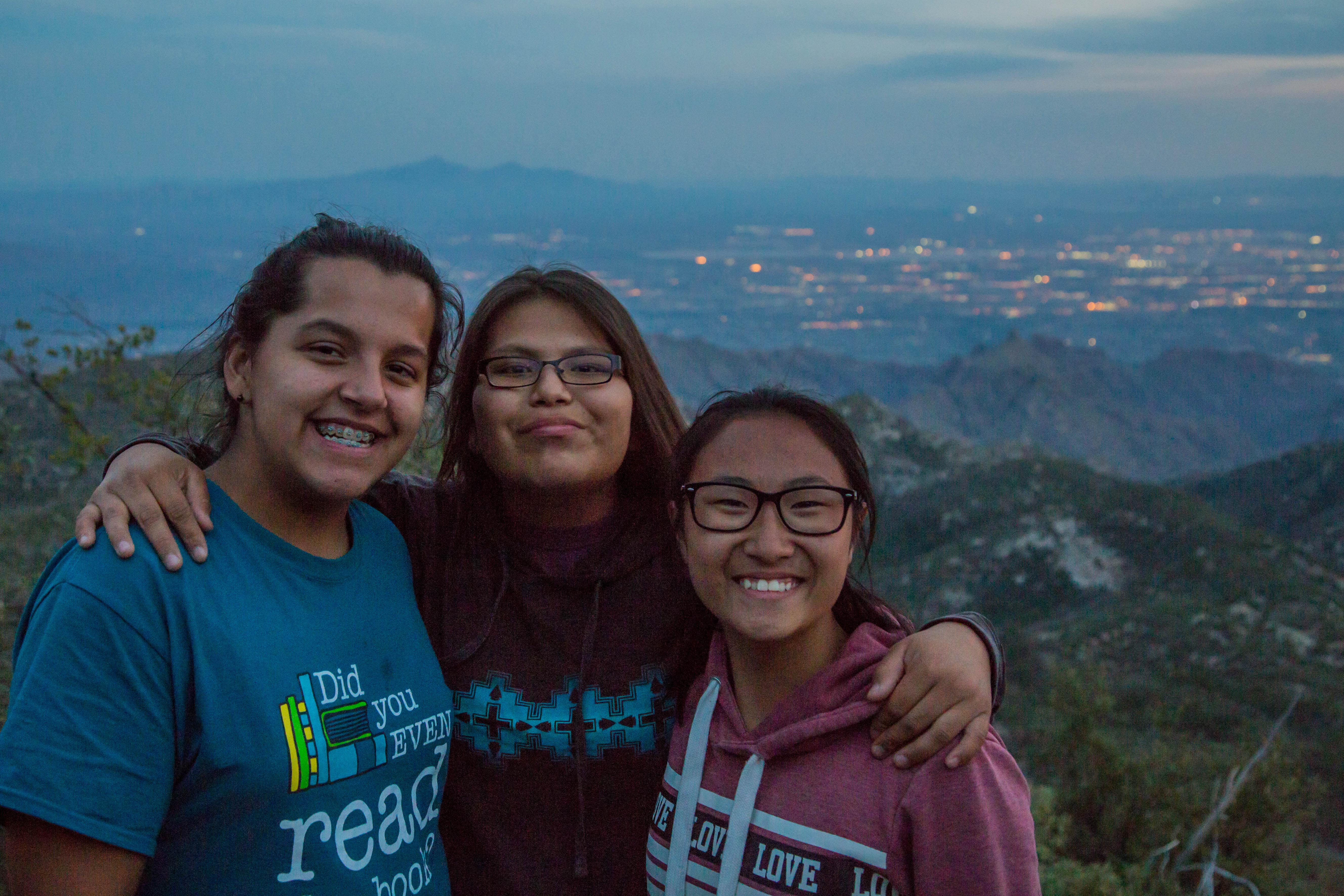 Three girls smile and pose for a photo with city lights and mountains in back