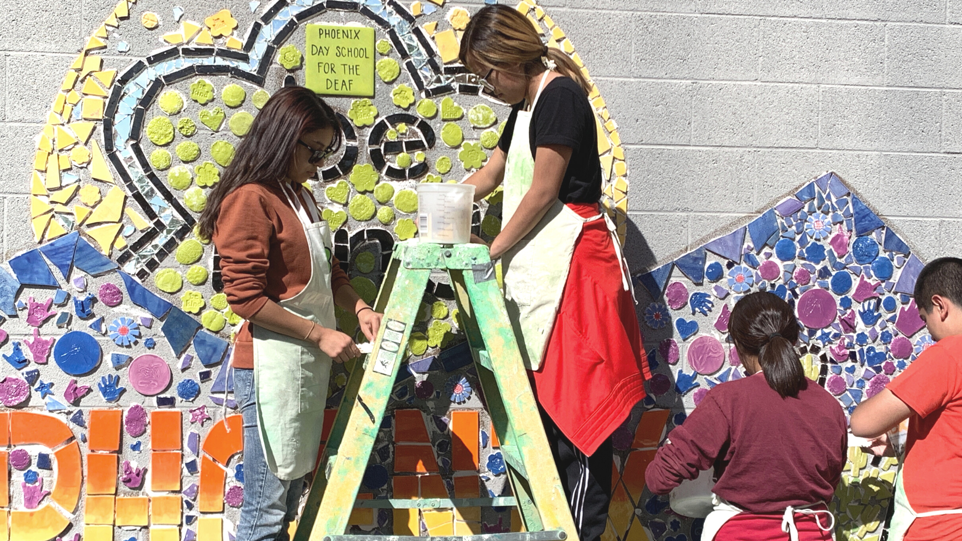Two students are seen standing on a ladder painting a wall