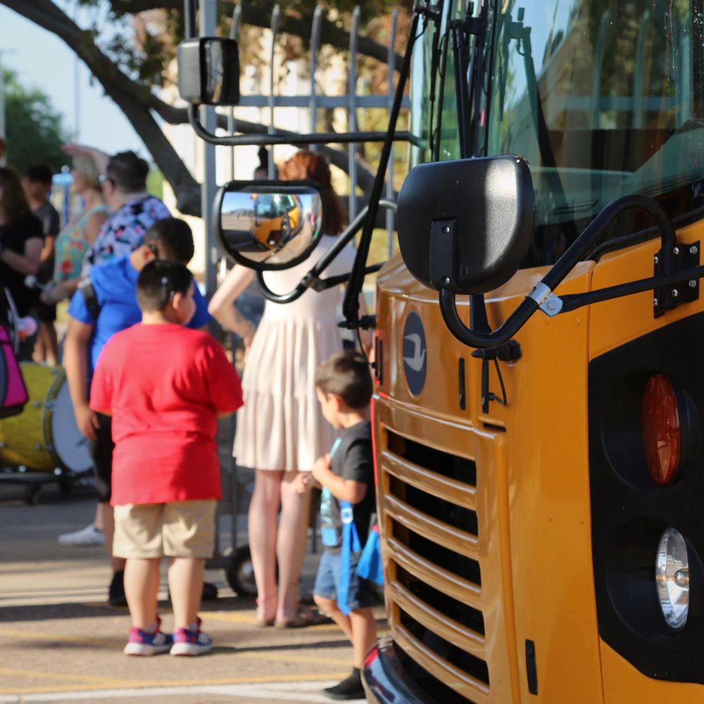 Children are gathered near the front gates of the PDSD campus next to a yellow bus.