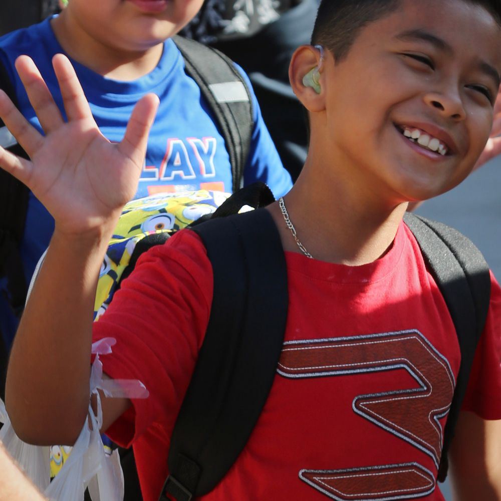 A young student wearing a red shirt and hearing aids smiles and waves at the camera.