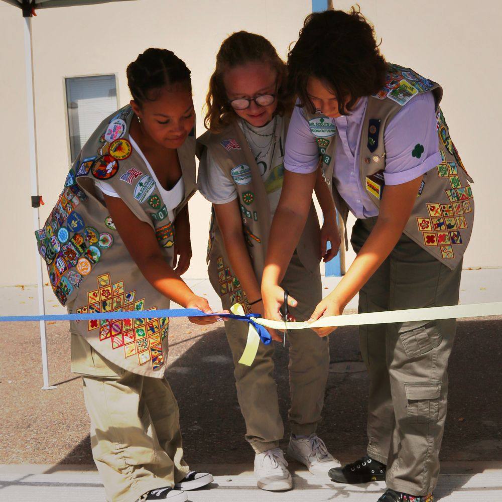 A photo of 3 girl scouts cutting a ribbon in their tan vests. The vests are covered in patches.