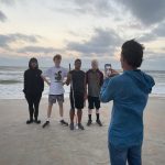 Photo of a person taking a photo of four students standing on the beach