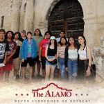 Image of a group of students standing in front of a large door with text that reads, "The Alamo Never Surrender Or Retreat."