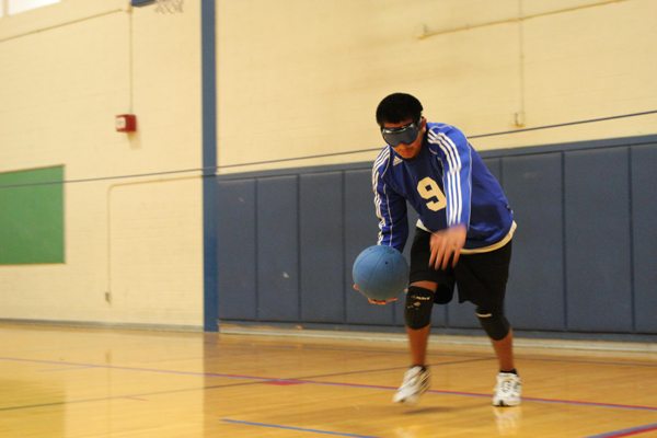 An ASB HS student playing Goalball is holding a Goalball at knee height and he appears ready to throw it during a game.