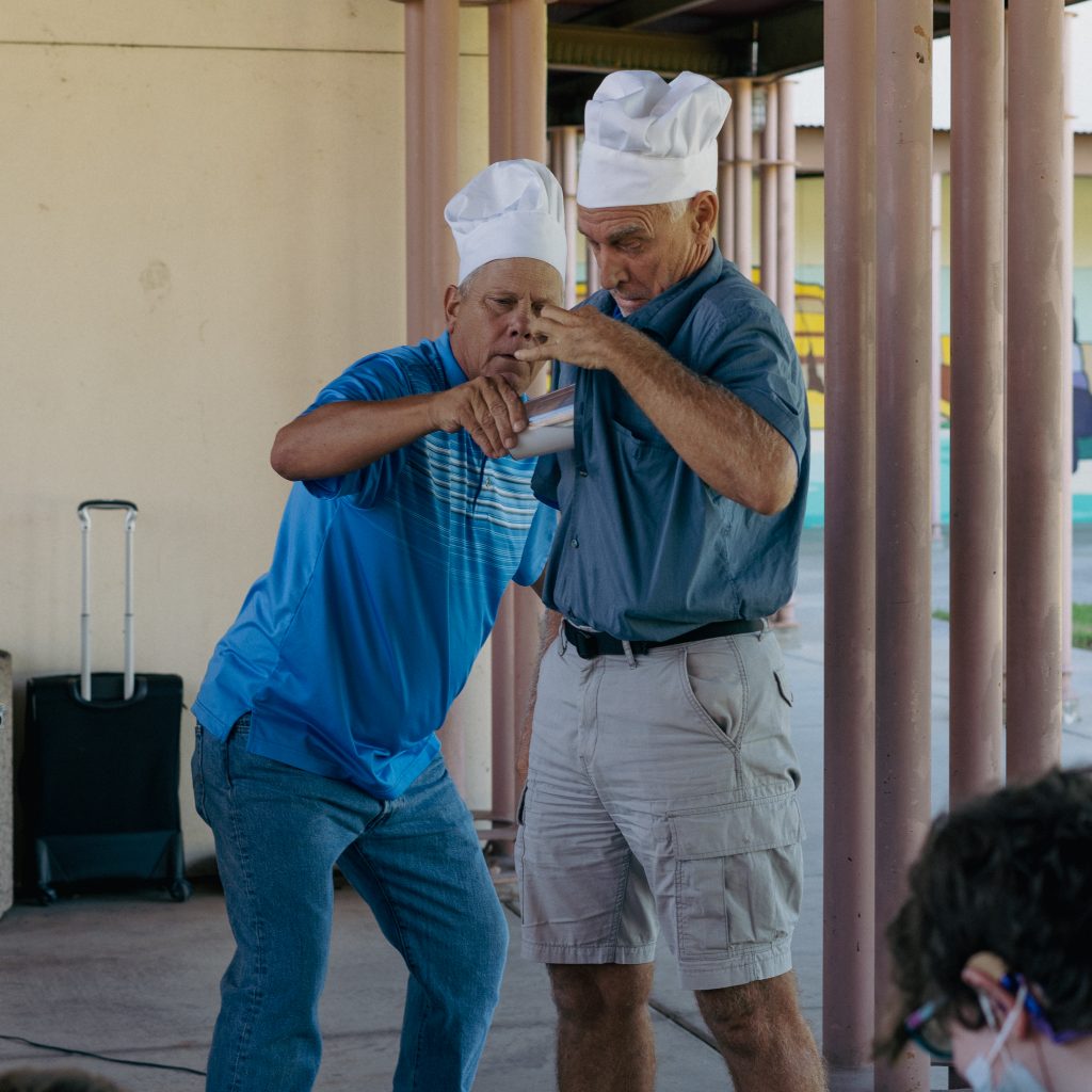 DJ Magic and A staff member wear white chef's hats and blue shirts. The staff member has a button-up shirt and DJ magic is pouring milk into his shirt. The staff member makes a disgusted face as he watches the milk pour into the shirt.
