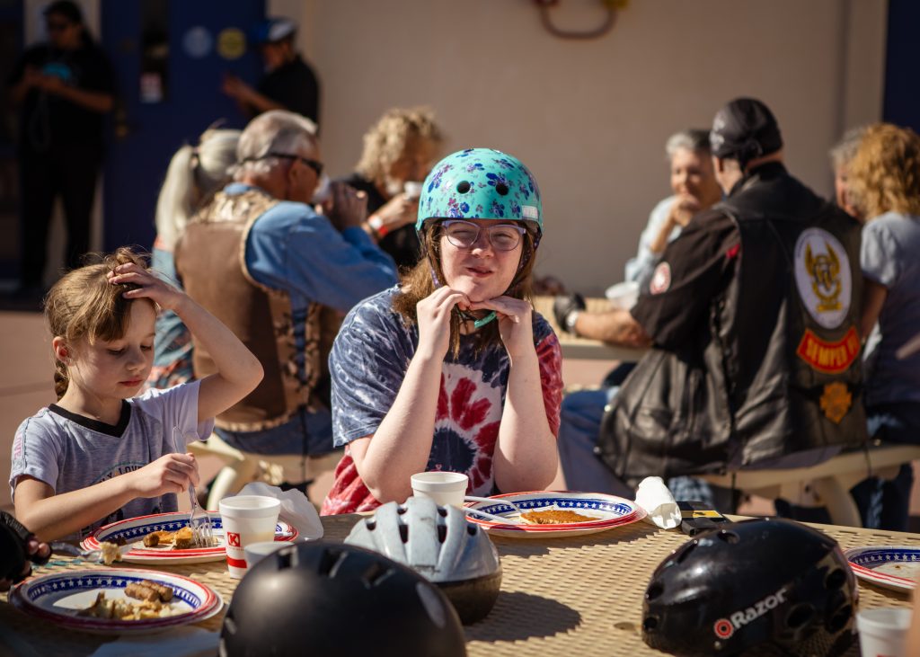 A student smiles with their hands under the chin posing for the camera. They are seated at a table, wearing a tye dye shirt and a teal blue helmet.
