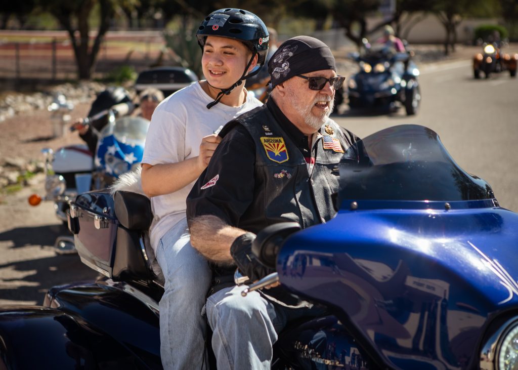 A student in a black helmet rides on the back of a blue motorcycle, holding on to the biker's leather vest.