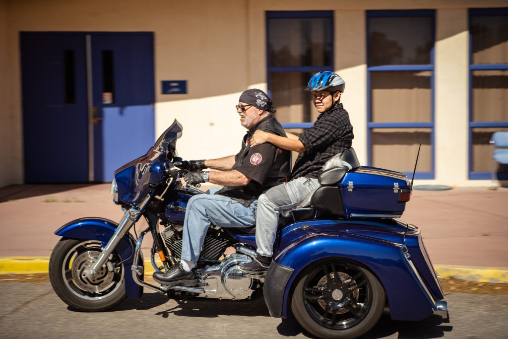 A student in a blue helmet smiles as they ride away on a blue tricycle motorcycle.