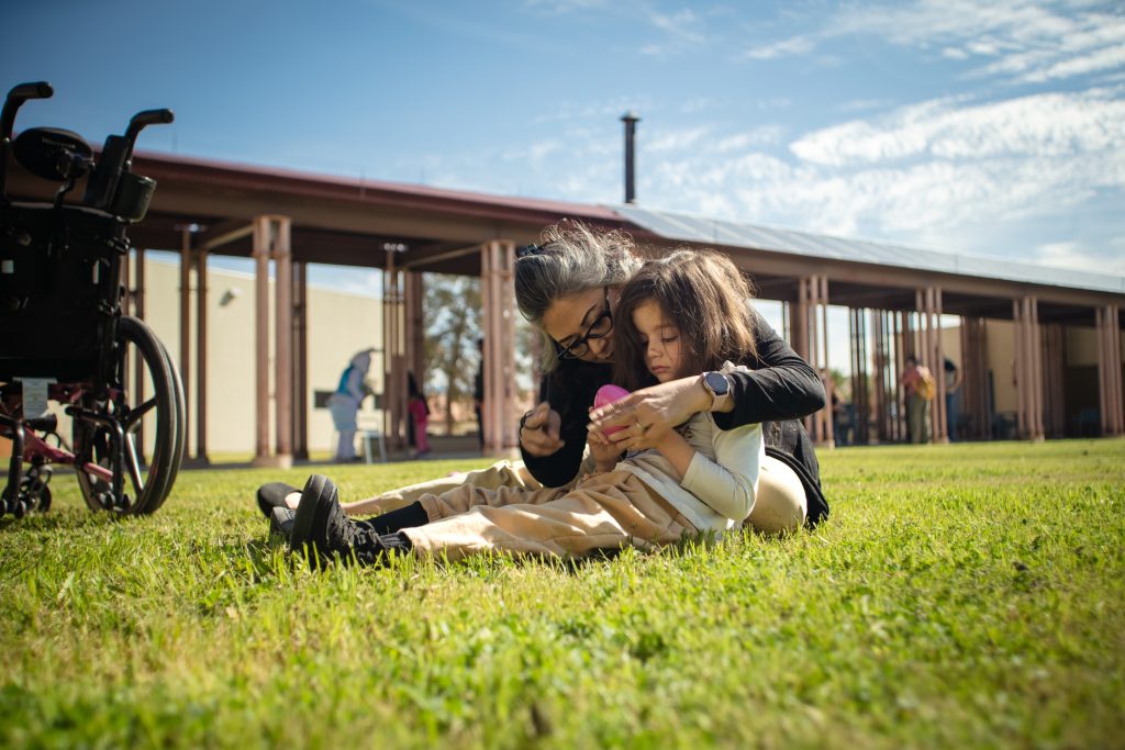 A student and staff member sit in the grass and enjoy a pink easter egg as the staff member signs "egg" in ASL