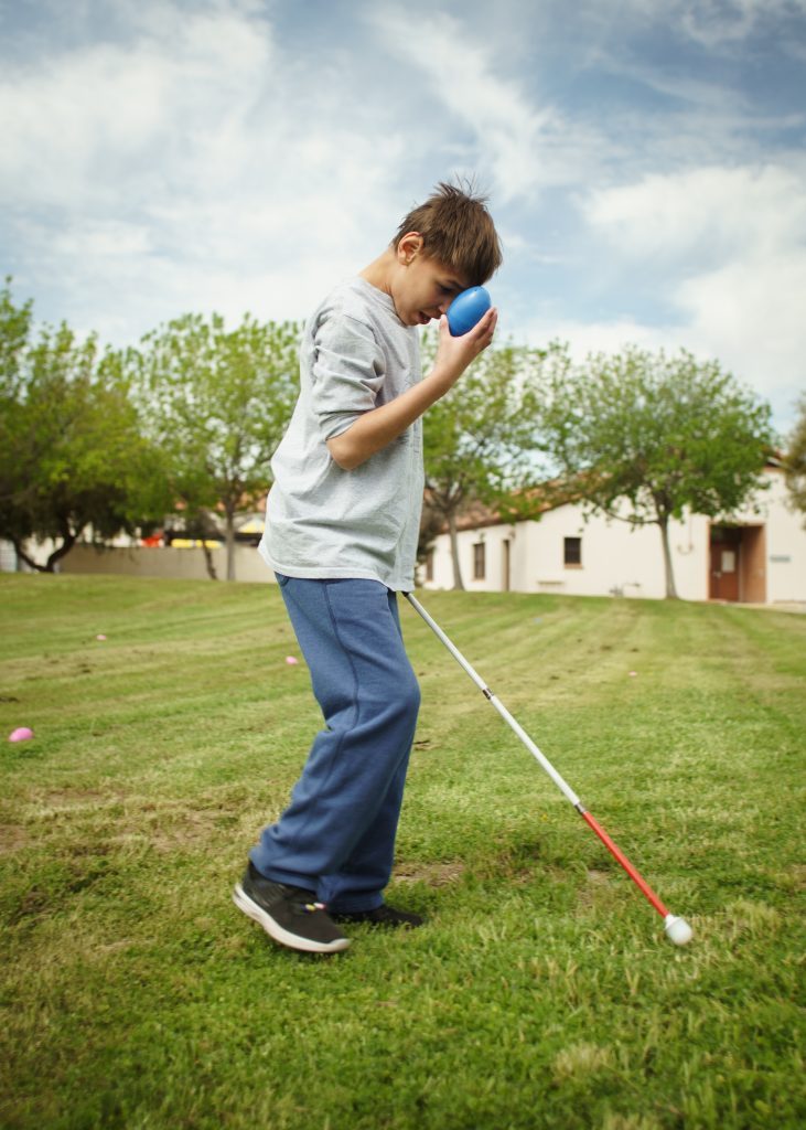 a visually imparied student finds a blue easter egg and smiles as they head across the grass with their white cane.