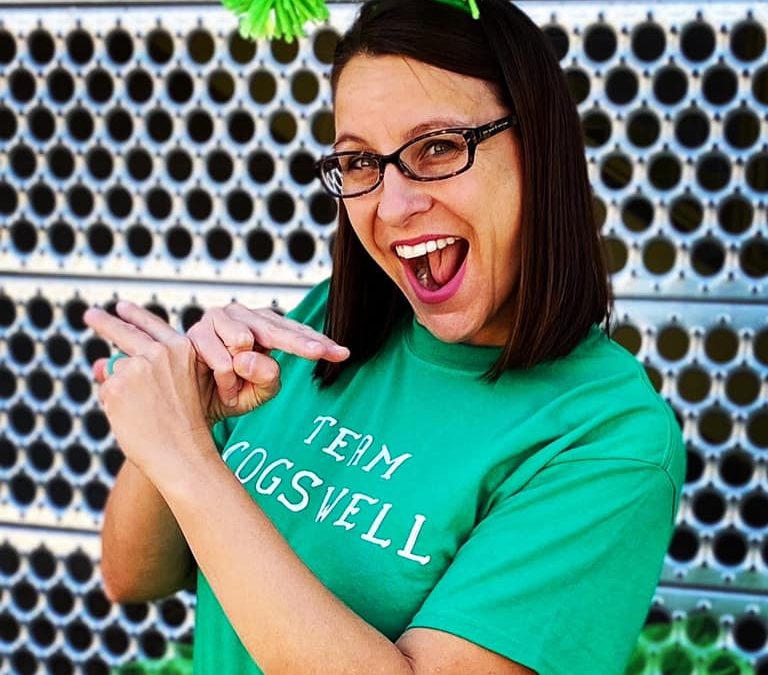 Jessica wearing a bright green headband with green yarn puffs on the top. She is wearing a green shirt that reads "Team Cogswell". She is smiling and holding her hands in the ASL sign Dragonfly.