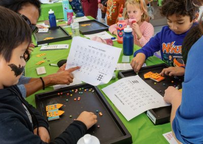 A photo of multiple students standing around a large outdoor table. Four of them are participating in an activity to strength their braille skills. The young students are using a braille template and colorful beads to help them form the letters of the braille alphabet.