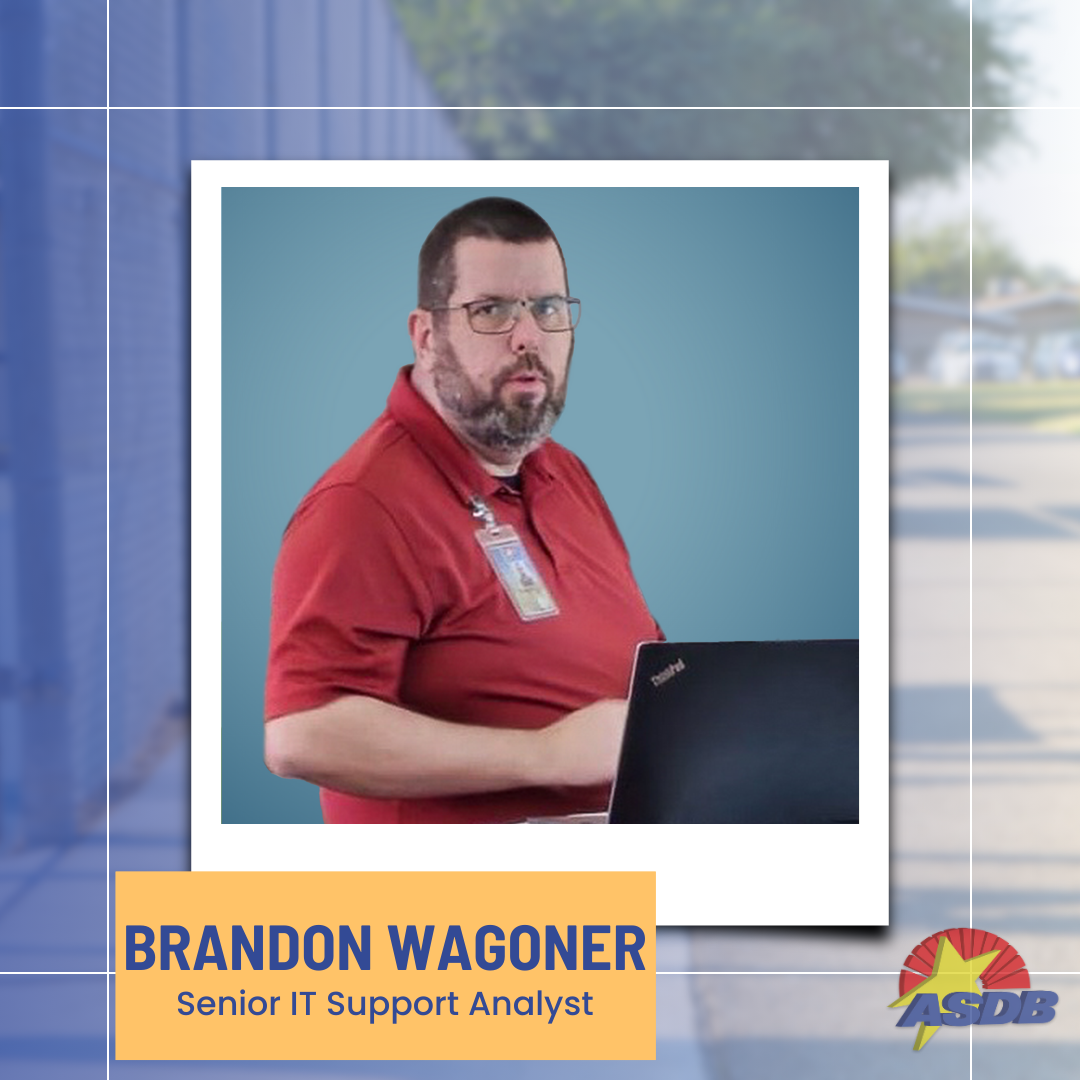 A portrait photo of Senior IT Support Analyst Brandon Wagoner. He has a buzzcut, a full beard, and is wearing a red polo shirt while standing in front of an open laptop.
