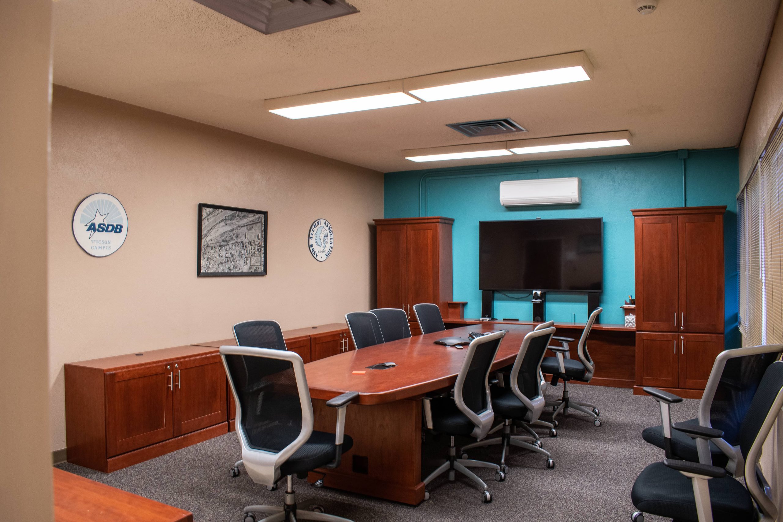 A wide photo of one of the conference rooms on ASDB campus. This particular conference room has an extended table and multiple office chairs. It also has rows of wooden cabinetry and a mounted flatscreen television.