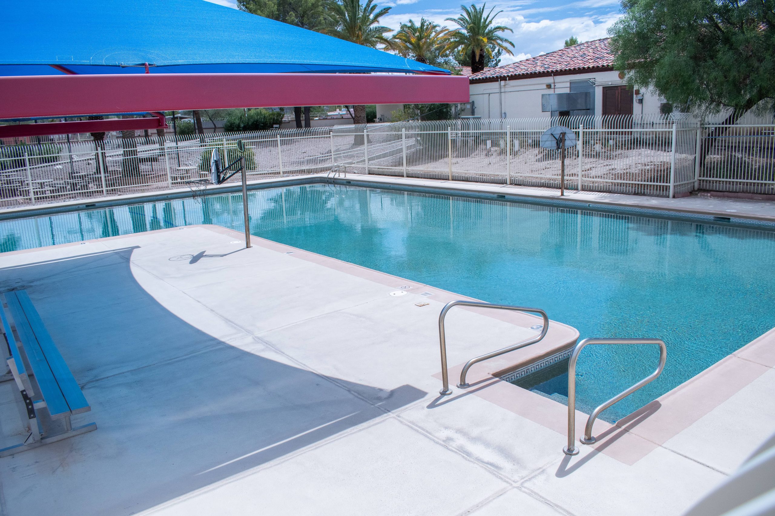 A wide photo of the swimming pool on ASDB campus. The pool has two entrances/exits, and vaguely resembles a right angle. Just beside the pool is a large, blue overhang shading the concrete walkway.